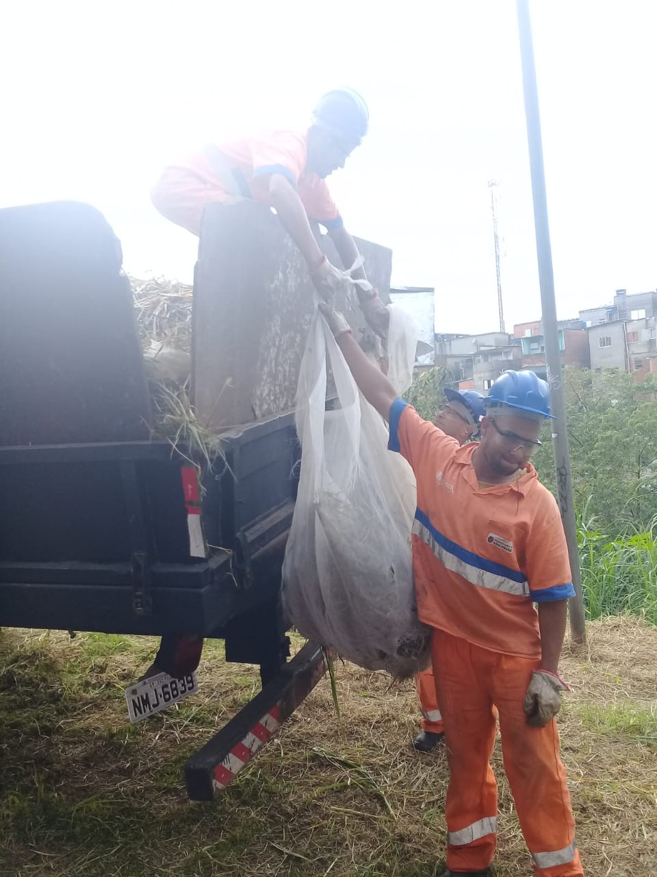Um funcionário abaixo entrega o saco de grama recolhida a um funcionário que está sobre um caminhão, ambos de uniforme laranja e capacete azul.